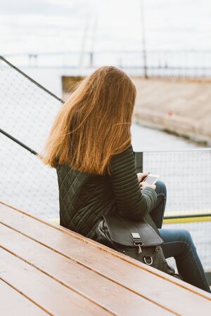 Unrecognizable woman with long hair sits with phone in her hand, her back to camera. Autumn or winter, a girl in jacket outdoors waiting for a call or sms, texting, verticalの写真素材