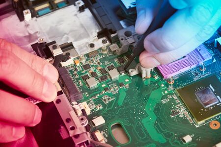 Man repairs computer. A service engineer in a shirt repairs a laptop, at a white Desk against a white wall. Computer motherboard background with blur neon multicolor glow. Hands close up.の写真素材