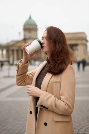 Beautiful serious stylish fashionable smart girl drinks coffee, goes walking down street of St....
