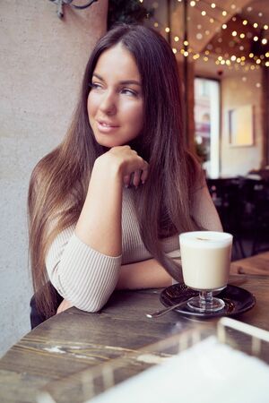 Beautiful girl sits in cafe in Christmas holidays, in background lights of garland. Brunette woman with long hair drinks cappuccino coffee, latte and looks out window, vertical, dark backdropの写真素材
