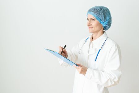 Female adult doctor stands on white background. Beautiful woman, thinking, smiling, writing. Clipboard in his hand, stethoscope around his neck .の写真素材