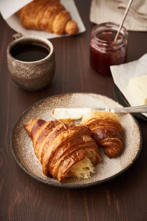 Morning breakfast with croissant on plate, cup of coffee, jam and butter. Dark brown table , morning...