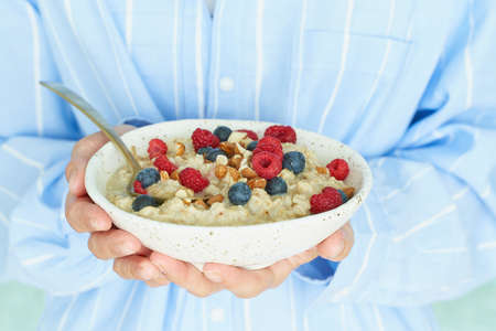 Faceless woman holds in hands breakfast, oatmeal porridge with berries and nuts, healthy food, proper nutrition. Sunshine, early morning, quiet home environment. Side viewの写真素材