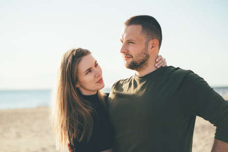 Beautiful woman full of love looking to handsome convinced man embracing her. Young adult couple standing on coast and huging each other. Millennial male and female posing on beach on sunny dayの写真素材