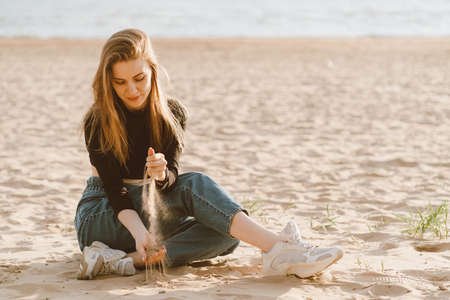 Full length beautiful woman sitting on beach at sunset in evening and pour sand. Slow life, social detox and self-immersion, single. Pretty female with long brown hair in casual clothes.の写真素材