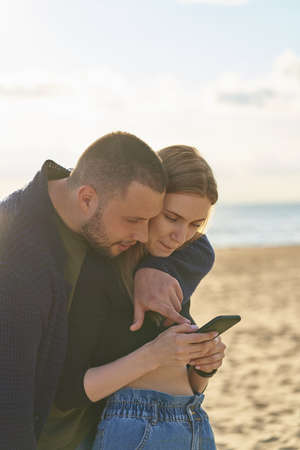 Young couple standing on sandy beach and looking at smartphone, man points with finger touching at mobile phone screen and selects photos for social networks. Walk of beautiful people in loveの写真素材