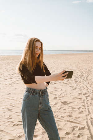 Beautiful woman taking selfie on ocean or sea coastline in sunny day. Pretty female with long hair, blonde takes photo on mobile phone on sandy beach in summer or autumnの写真素材