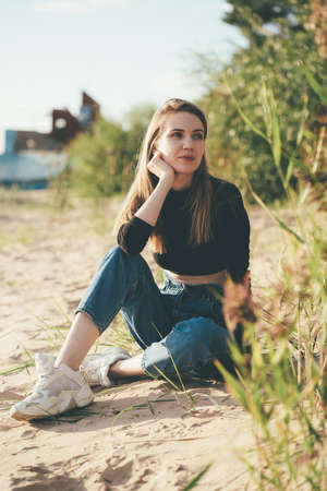 Full length beautiful pensive woman sitting on beach at sunset in evening. Pretty female with long...