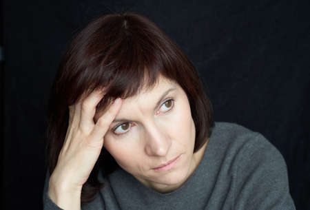 Mature, middle-aged woman looks anxiously away, touching forehead and hair with handの写真素材
