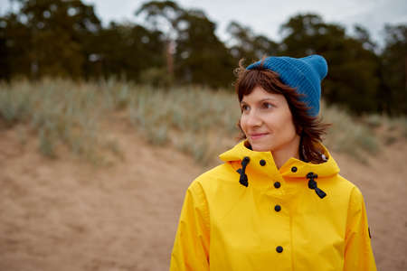 One with nature. Mature beautiful woman looking to forest in bright yellow jacket and blue hat on...