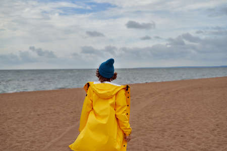 Back view of woman in bright yellow cloak and red hat on shore of North sea on cold autumn day. Mood Scandinavian weather, deserted beachの写真素材