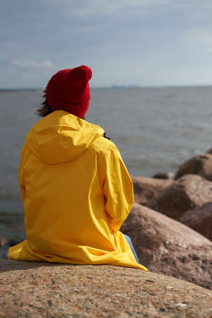 Back view of woman waiting and sitting in bright yellow cloak and red hat on stone near shore of North sea on cold autumn day. Mood Scandinavian weather, deserted beach, coast.の写真素材