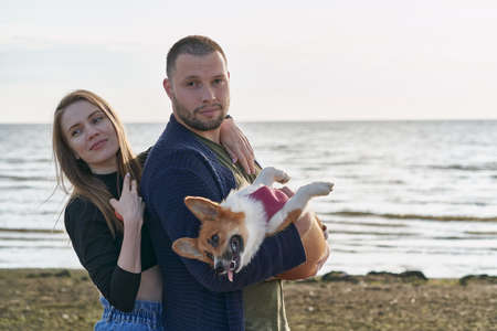 Young happy couple with dog standing on beach. Beautiful girl and guy and Corgi puppy having fun....