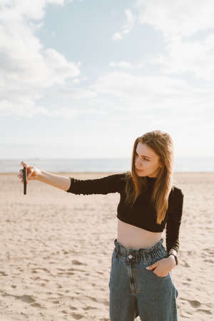 Pretty female with long hair, blonde takes photo on mobile phone on sandy beach in summer or autumn. Beautiful woman in jeans and black topの写真素材