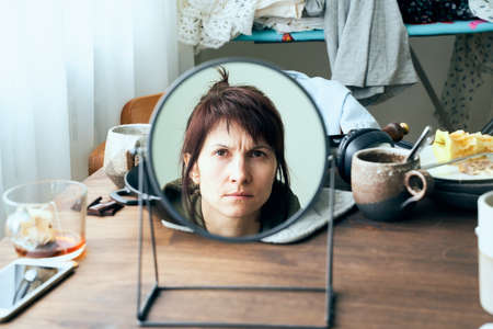 Woman looks at herself in mirror against background of mess, dirty dishes, piles of clothesの写真素材