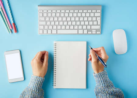 Faceless woman writing in notepad. Blank white notepad. Mobile phone with blank white screen display. Bright blue modern desk with smartphone. Top view. Copy space, mock up.の写真素材