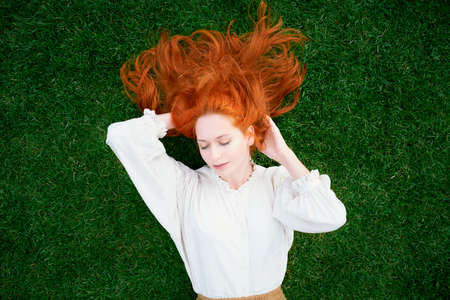 Beautiful girl with red hair lying on green grass with closed eyesの写真素材