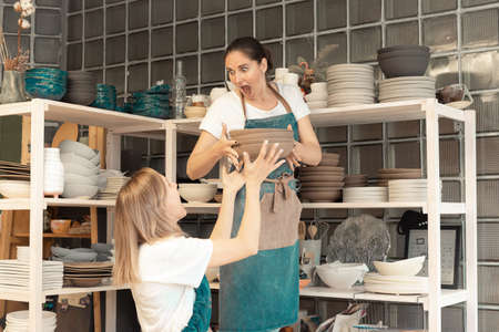 Two female entrepreneurs in ceramic studio pass dishes to each other. Woman joking and pretending that dishes fallの写真素材