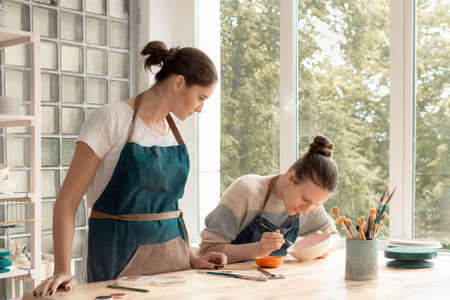 Woman making ceramic pottery. Attractive skilled young lady in apron standing at table and teaching student. Concept for workshop and master classの写真素材
