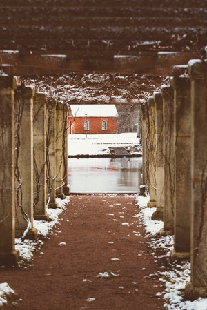Brick red house at end of snow-covered path, view through wooden frame, surrounded by trees and...