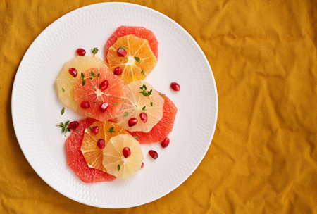 Bright mix of citrus fruits. Salad of mix sliced round slices of red and white grapefruit sprinkled with pomegranate seeds on white plate on yellow linen. Copy spaceの写真素材