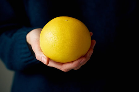 Close-up of womans hand holding whole white yellow bright citrus fruit grapefruit on dark...