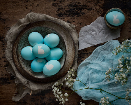 Unusual Easter on dark old background. Ceramic brown bowl with blue eggs with rabbit. Darkness, rays of sunlight shine on eggs.の写真素材
