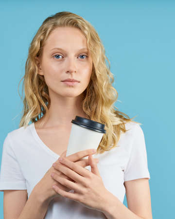 Portrait of young student girl with cup of coffee on color neutral tone blue wall. Pretty serious...