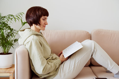 Mature woman reading book sitting on sofa, thinking alone. Digital detox concept, free time for yourself on weekends Saturday and Sunday. Self-immersion, self discovery. Relaxing and reflectionの写真素材