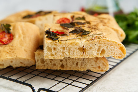 Slice of chopped focaccia, pizza, cut flat bread with vegetables on grid, tradition italian cuisine. Homemade artisan flatbread with tomatoes, parsley on white pastel table. Side view, close upの写真素材