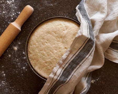 Raw yeast dough resting and rising in large metal bowl covering with linen towel on dark background....