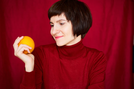 Smiling mature brunette woman looking at orange fruit in hand, beautiful middle-aged female in bright red sweater on bright colorful dark red background. Generation X, 40s, luxury interiorの写真素材
