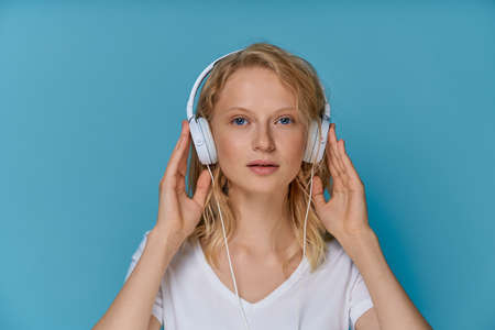 Closeup portrait of young woman listening music via headphones on color bright blue tone wall. Pretty serious clever blonde woman with curly hair looking at cameraの写真素材