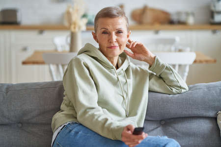 Movie time. Portrait of beautiful middle aged woman with remote control in hand watching tv while sitting and relaxing on sofa, spending leisure time at home. Selective focus on senior femaleの写真素材