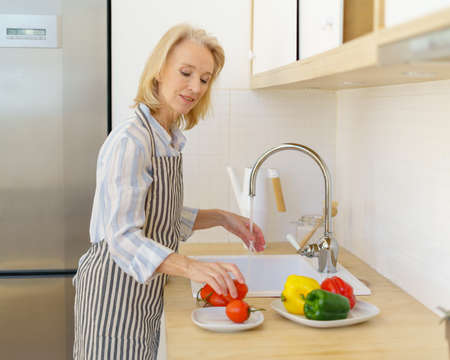 Happy senior woman grandmother in apron washing fresh vegetables for salad while enjoying cooking in modern light kitchen at home. Smiling elderly woman preparing healthy food for familyの写真素材