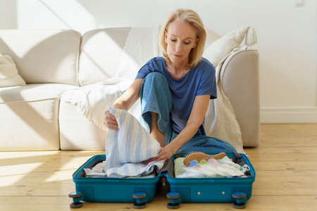 Thoughtful mature 60s woman packing suitcase, preparing luggage for travel trip while sitting on wooden floor at home. Senior lady getting ready for summer vacation. Traveling in retirement conceptの写真素材