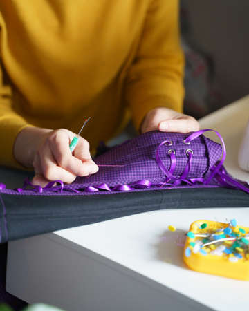 Woman seamstress or tailor using sewing hook while making purple corset with lacing, working at her creative workplace in atelier or home with colored pins on table. Lingerie manufacture conceptの写真素材
