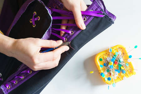 Cropped shot of female seamstress or tailor using scissors while sewing purple corset with lacing at her workplace in atelier or home, sitting at table with colored pins. Lingerie tailoring processの写真素材