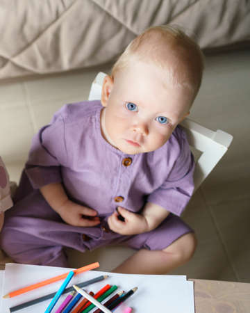 Cute blue eyed little baby boy with golden hair in linen romper looking at camera while sitting at desk and drawing with colorful pencils, view from above. Early childhood developmentの写真素材