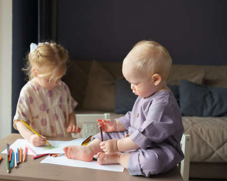 Cute baby boy with golden hair sitting on table, playing with colorful pencils while his older...