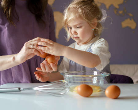 Adorable little girl with blonde hair helping mom to cook pie, mother showing daughter how to break...