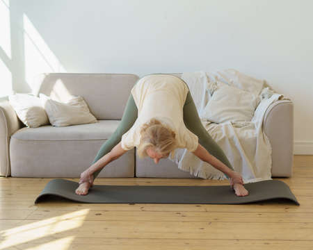 Active senior 60s woman in sports wear practicing downward facing dog yoga pose while exercising at home. Mature flexible lady doing wide legged forward bend or Prasarita Padottanasana postureの写真素材