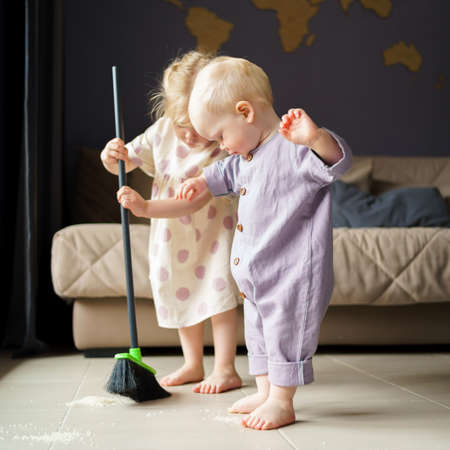 Cute little children infant boy and toddler girl in linen clothes sweeping floor in room with broom, helping mom with house chore at home. Baby boy brother and older sister spending time togetherの写真素材
