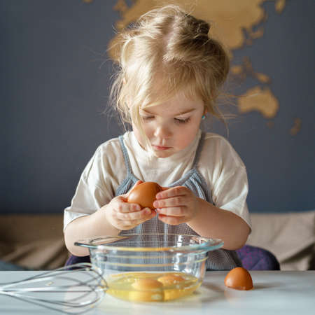 Focused little girl with blonde hair breaking fresh eggs into bowl for omelet or pie while standing...