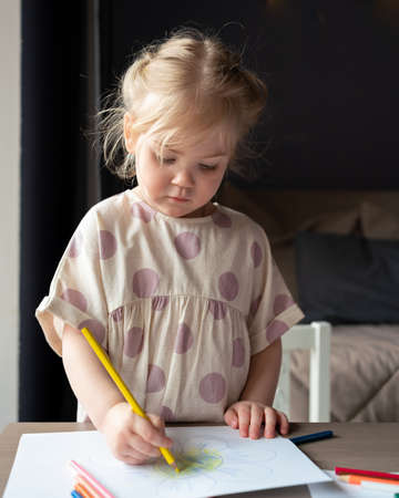Charming little preschool child girl with golden hair in linen dress drawing on piece of paper with colorful pencils while playing at home, vertical shot. Creative learning and development for kidsの写真素材
