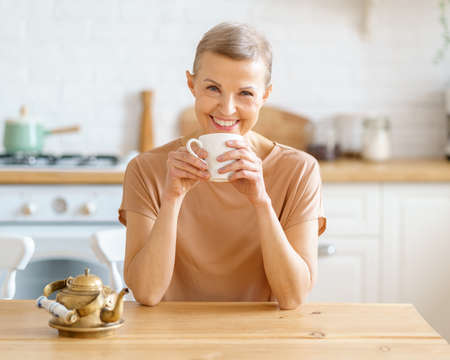 Morning drink. Beautiful senior woman looking to camera and drinking coffee and thinking about something while sitting at wooden table in kitchen, selective focus on female. Mature people lifestyleの写真素材