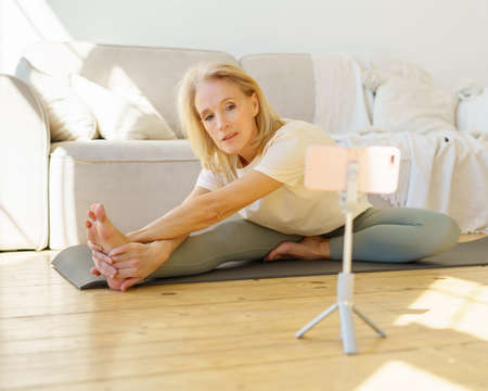 Flexible retired woman exercising on yoga mat in living room, watching online class on smartphone,...