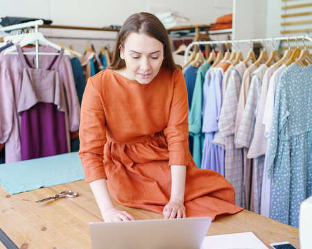 Working on new collection. Young successful business woman fashion designer sitting on table and works on laptop at her own workshop, female dressmaker or seamstress creating clothing design onlineの写真素材
