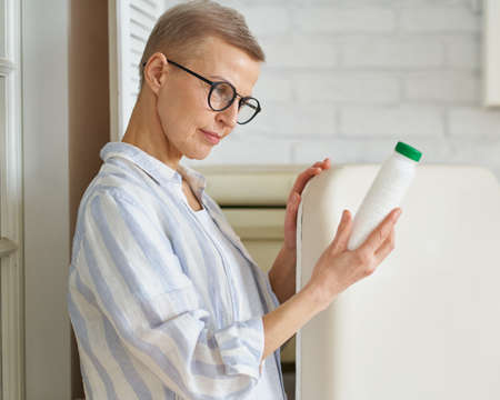 Beautiful senior woman wearing glasses looking at bottle with yogurt, check shelf life and ingridients of product in kitchen near opened refrigerator. Mature people and healthy eating conceptの写真素材