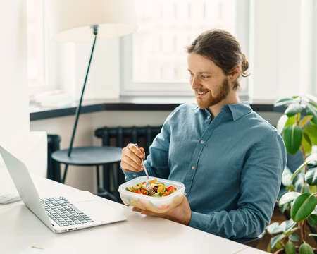 Satisfied male employee eating vegetarian food, pasta with vegetables, holding plastic container with vegan meal and watching funny video on laptop while having break in office, enjoying lunch at workの写真素材